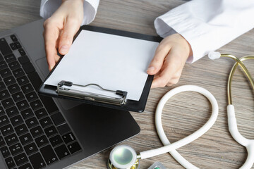 Telehealth. Doctor with clipboard, laptop and stethoscope at wooden table, closeup