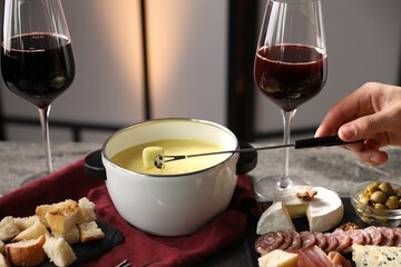 Woman dipping piece of cheese into fondue pot (caquelon) with melted cheese at table indoors,...