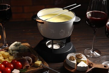 Fondue pot (caquelon) with melted cheese, wine and snacks on wooden table, closeup