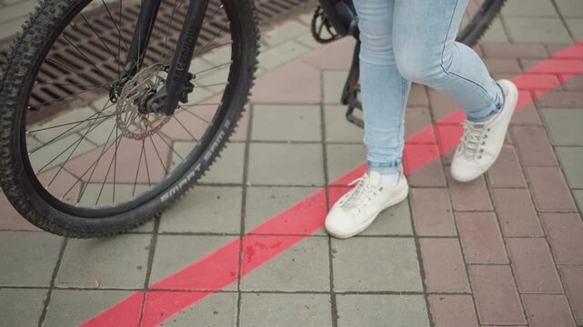 woman beside bicycle on red curb, jeans and white sneakers step slowly across tiled pavement near bike wheel and pedal, moody urban atmosphere conveying solitary commuter routine and casual street