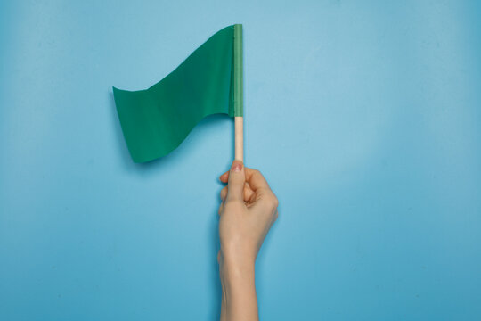 Hand holding a green flag against a blue background. Concept of permission, approval, safety, positive signal, start or go ahead symbol with copy space.