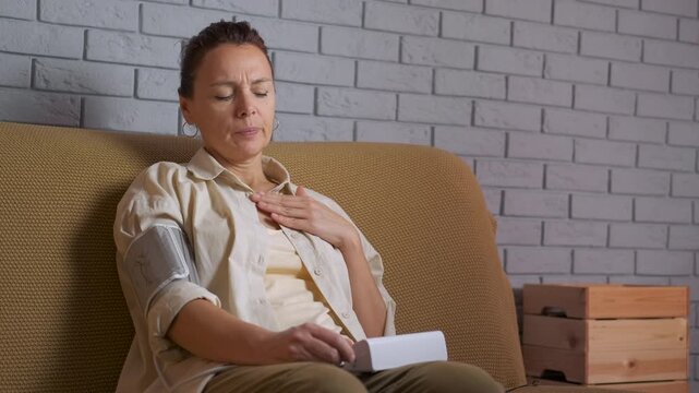 Woman measuring blood pressure having chest pain at home. Stressed mature woman measuring her arterial blood pressure with an electronic tonometer, feeling unwell and experiencing chest pain while
