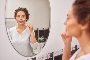 A woman with curly hair is using a facial tool to apply a skin care product while gazing at her reflection in a bathroom mirror during her morning routine.