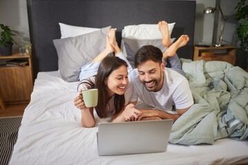 Two people are lying together in bed, engaged in a joyful activity on a laptop, while sharing a warm drink, creating a relaxed and intimate atmosphere in their bedroom.