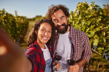 Two smiling individuals hold glasses of red wine, standing among lush green grapevines. They are capturing a joyful moment at their family vineyard under a clear blue sky.