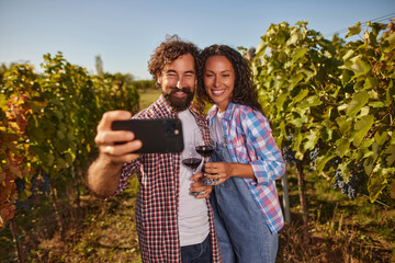 A happy couple stands in a vineyard, holding glasses of red wine as they take a selfie together. Lush grapevines surround them under a clear blue sky.