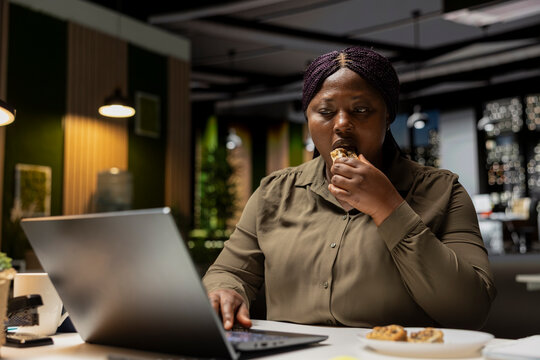 Young businesswoman spends night time at the office eating unhealthy food, dealing with a stress eating disorder due to hard work after hours. Emotional compulsive overeating at the firm.