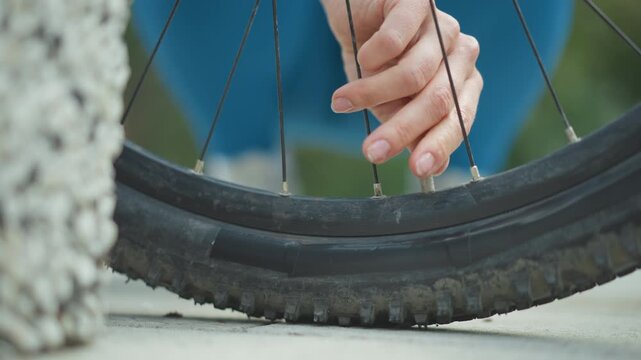 closeup bicycle flat tire repair by womans hand, urban sidewalk, deflated knobby tire against concrete, fingers checking valve and spoke, patient slowmotion maintenance, soft morning light,