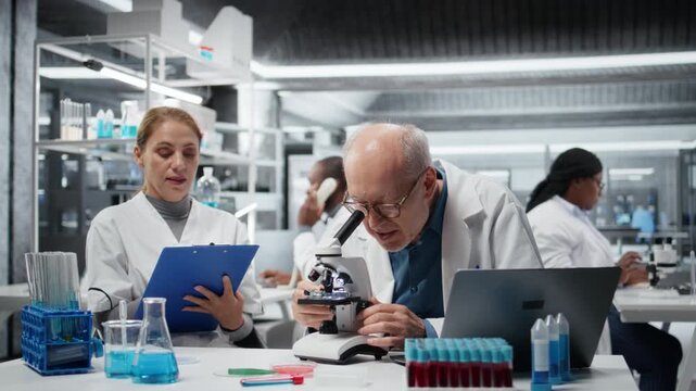 Laboratory technician examining biopsy slide while colleague documents diagnostic findings on clipboard. Elderly man identifies disease markers in clinical lab with coworker taking notes, camera A