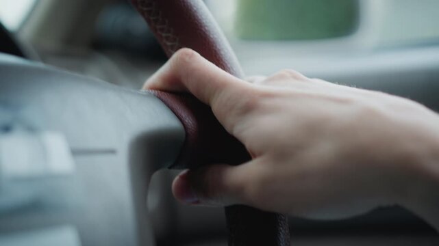 Tight shot hand on steering wheel inside car, firm grip on leather rim, dashboard and window blurred, outside greenery hinting suburban drive, driver steering with concentration, parent chauffeur