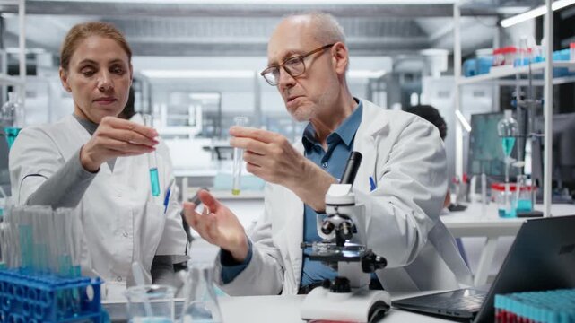 Lab team leader teaching intern to analyze liquid polymer properties in test tube. Senior scientist mentoring trainee to test new synthetic material viscosity in laboratory, camera B