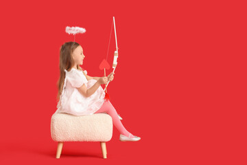 Fototapeta premium Cute little girl dressed as cupid with bow and arrow sitting on pouf against red background. Valentine's Day celebration