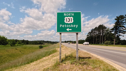 Northbound US-131 Highway Sign Pointing to Petoskey, Michigan. Set against a blue sky with fluffy white clouds, situated on the side of the road exit ramp. A major transportation corridor in Michigan.
