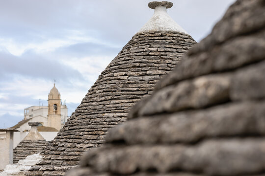 conical limestone roofs of traditional trulli houses in alberobello, apulia, italy, featuring stacked stones, a cloudy sky, and a historic church bell tower.
