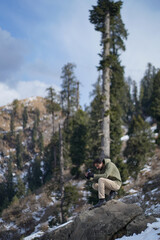 Obraz premium Man in olive hoodie crouched on a rock using a smartphone to photograph a vast, snowy mountain forest under a blue sky.