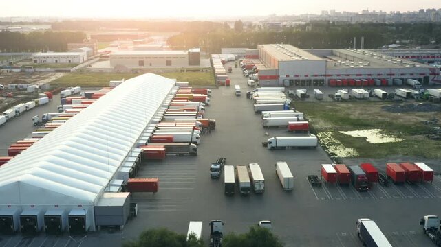 Aerial View: Large Logistics Distribution Hub with Semi Trucks at Loading Docks | Modern Warehouse Complex with Cargo Trailers Parked at Sunset | Industrial Supply Chain Operations