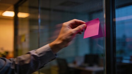 A business person's hand quickly sticking a blank pink sticky note on a transparent glass wall in a modern office at dusk