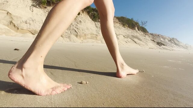 Close up of a woman walking barefoot on sandy beach near rocks and dunes. Steps in wet sand. Summer vacation, relaxation, freedom, seaside lifestyle, and nature journey