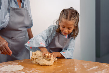 Little girl kneading dough on kitchen table while baking with grandmother at home wearing aprons and enjoying fun family cooking activity together