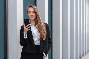 Confident young businesswoman in black suit using smartphone near modern building with professional and stylish appearance during daytime