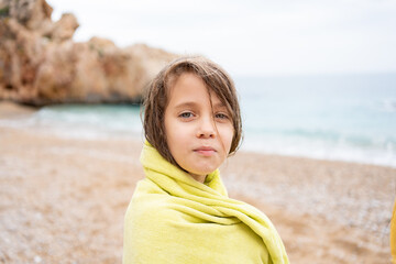 Happy girl wrapped in yellow towel standing near sea on sandy beach after swimming. Cute child is enjoying vacation by seaside