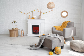 Naklejka premium Interior of stylish room with modern fireplace, armchair, table and autumn decorations near white brick wall