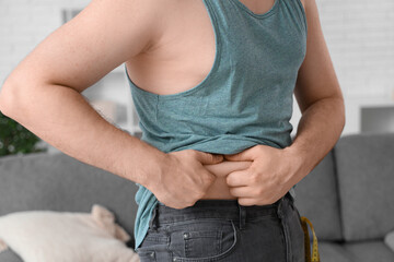 Young overweight man touching fat on his belly at home, closeup. Weight gain concept