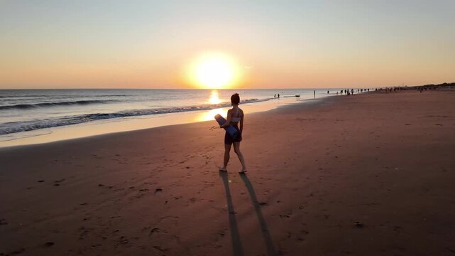 Athletic young woman walking on the beach with a yoga mat at sunset