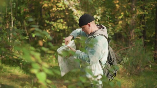 white hiker reading map in forest, backpacked navigator unfolds paper route between sunlit trees, scanning bearings and adjusting beanie while planning trail and checking foliage for landmarks, quiet