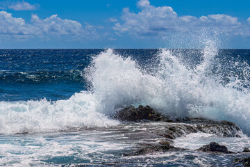 Fototapeta premium Wave breaking on the eastern coast of Hawaii's Big Island. Foam, volcanic rocks in the foreground. Blue Pacific Ocean, cloudy sky in the background. 