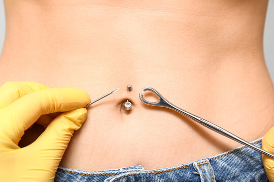 Professional doing piercing of young woman's navel on grey background, closeup