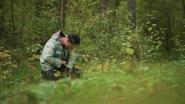 caucasian backpacker repairing strap in forest, kneeling behind low grass, adjusting buckle and cord, using hands to tighten gear, focused posture, misty green background, resourceful mood, rugged