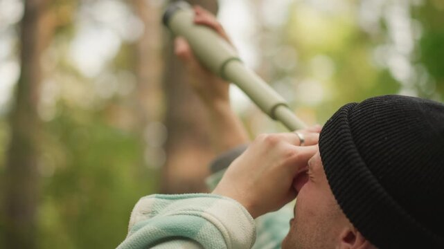 hiker in beanie peers through telescope into treetop, amateur birdwatcher scans canopy with olive spyglass, steady hands, dappled sunlight through pines, quiet curiosity and outdoor exploration mood