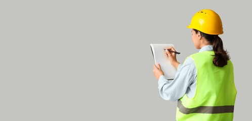 Young female construction worker writing in notebook on grey background with space for text