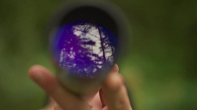 purple lens reflecting forest through monocular, hand holding tube against blurred green bokeh, closeup of trees mirror in glass, dreamy outdoor mood with subtle light and shade, nature photographer