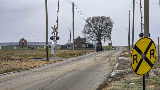 A dirt road runs along train tracks as a horse and buggie tracks on road. Utility poles line the road. A farmhouse is visible in the background, with solar panels nearby and overcast skies above.