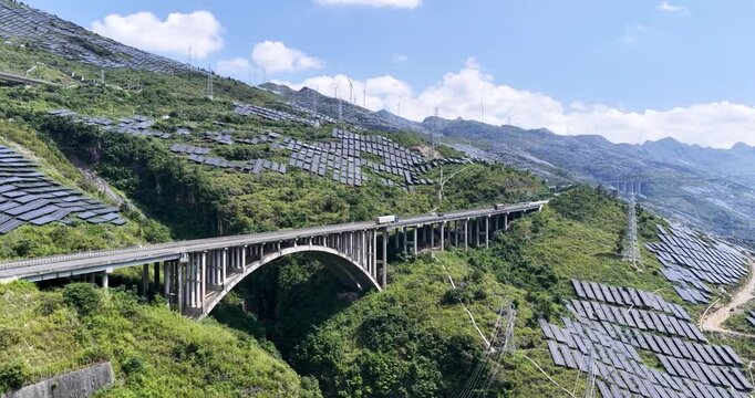 Aerial video of a highway crossing mountainous solar farms, with photovoltaic panels covering slopes and wind turbines in the distance under clear blue sky.