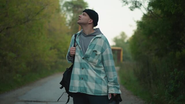 white male photographer walking on road, glancing up at canopy, holding camera and tripod, shoulder bag and beanie, soft golden light through trees, creative scouting and nature observation, content