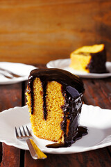 A slice of Brazilian carrot cake with a glistening chocolate glaze, served on a white plate atop a rustic wooden table