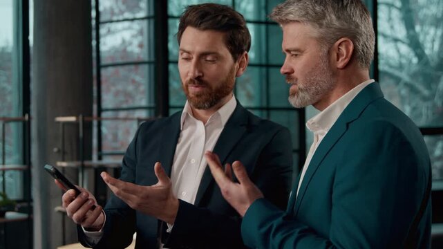 Two Caucasian businessmen discussing business idea in office using smartphone professional males men guys manager consultant showing mobile phone client salesman explaining benefits negotiating deal