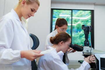 Fototapeta premium Team of female scientists collaborating in digital pathology lab. Young researcher adjusting microscope while colleague observes with tablet for medical diagnosis.