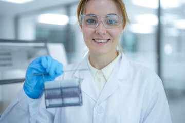 Fototapeta premium Portrait of a smiling female scientist holding a rack of microscope slides near an automated staining machine. Professional researcher preparing tissue samples for medical pathology diagnosis.