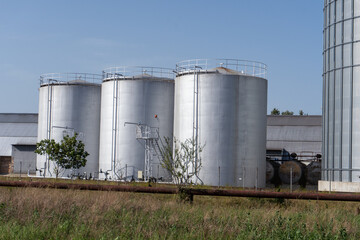 Obraz premium Three silver storage tanks stand at a facility surrounded by fields and buildings on a clear day