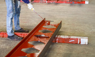 Worker is painting metal beam with red paint in industrial setting, showcasing construction process. beam is supported by wooden blocks on concrete floor