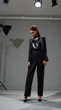 Fashion model in a sleek black outfit poses confidently under studio lights. Low-angle shot emphasizes elegance, perfect for a fashion video concept.