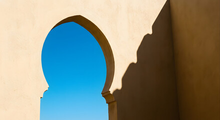 Minimalist moroccan architecture featuring a traditional arched portal against a clear blue sky with sharp sunlight casting a deep geometric shadow on a textured beige plaster wall