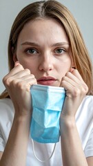 Young woman holding a blue surgical mask to her face, gazing intently forward against a neutral background