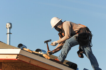 Construction worker using a roofing hammer on the roof structure of a wooden house under construction, representing exterior finishing work and active housing construction operations