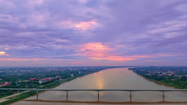 A wide aerial view of Kong river with a long friendship bridge Thai and Loas stretching across the water under a dramatic, cloud filled sunrise sky. Soft light breaks through the clouds