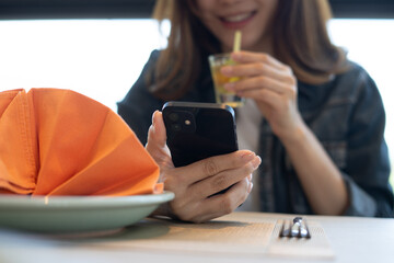 Happy young asian woman using mobile phone and drinking juice in restaurant, cafe interior. Smiling woman using smartphone for social media network, surfing internet, close up